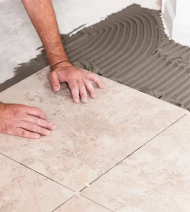 Hands set a large beige floor tile into fresh adhesive.
